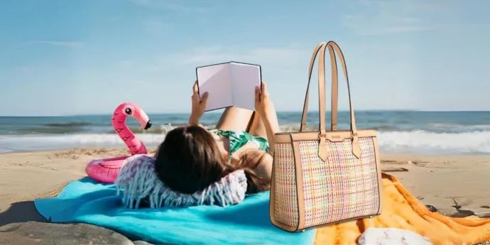 Bolso shopper Cala, uno de los bolsos de mujer de verano que son tendencia en 2024 sobre una toalla en la playa con una mujer leyendo.